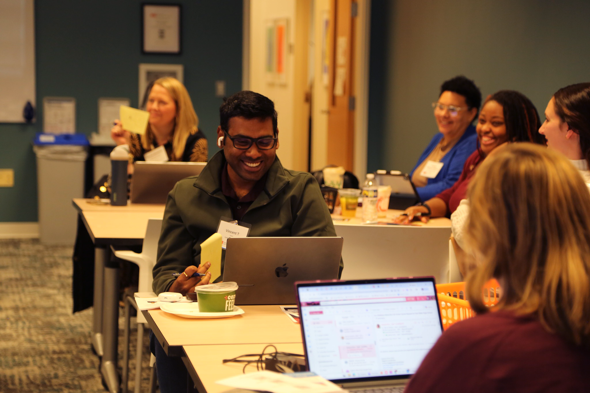A man smiles within a crowd of people sitting at tables in front of laptops