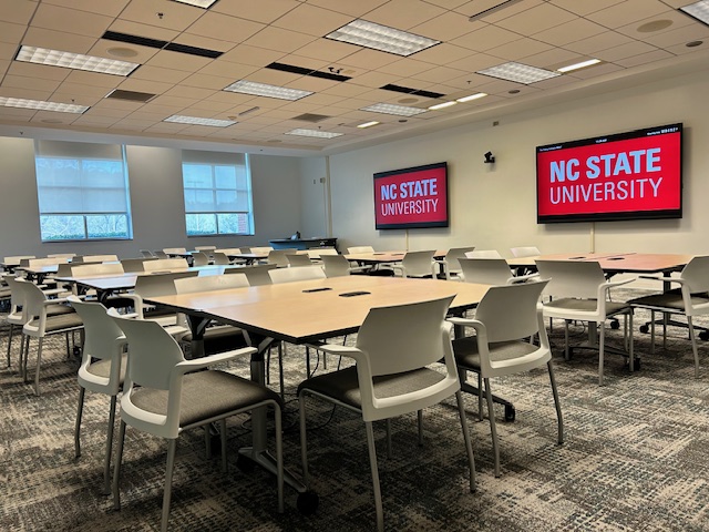 A meeting space with TVs on the wall and square tables with chairs