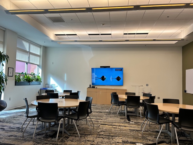 A meeting space with a tv on the wall and round tables with chairs