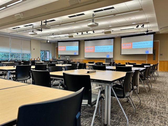 A large meeting space with projection screens on the wall and moveable square tables with chairs
