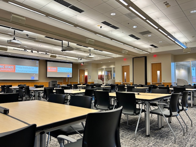 A large meeting space with projection screens on the wall and moveable square tables with chairs