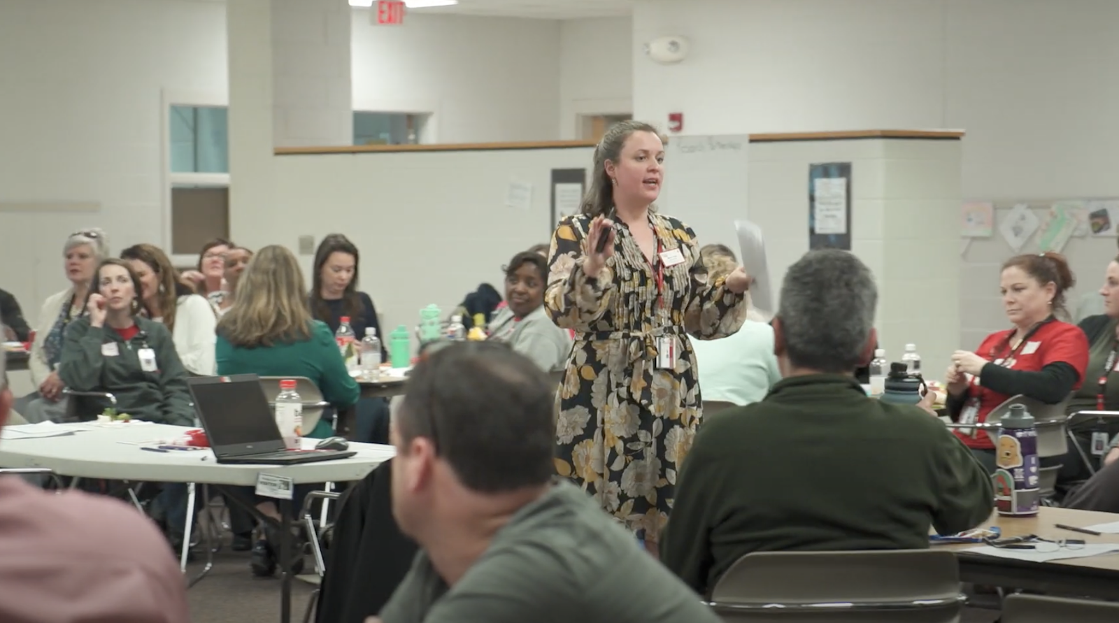 A woman with long brown hair and a dress speaks to a room full of teachers sitting at various tables