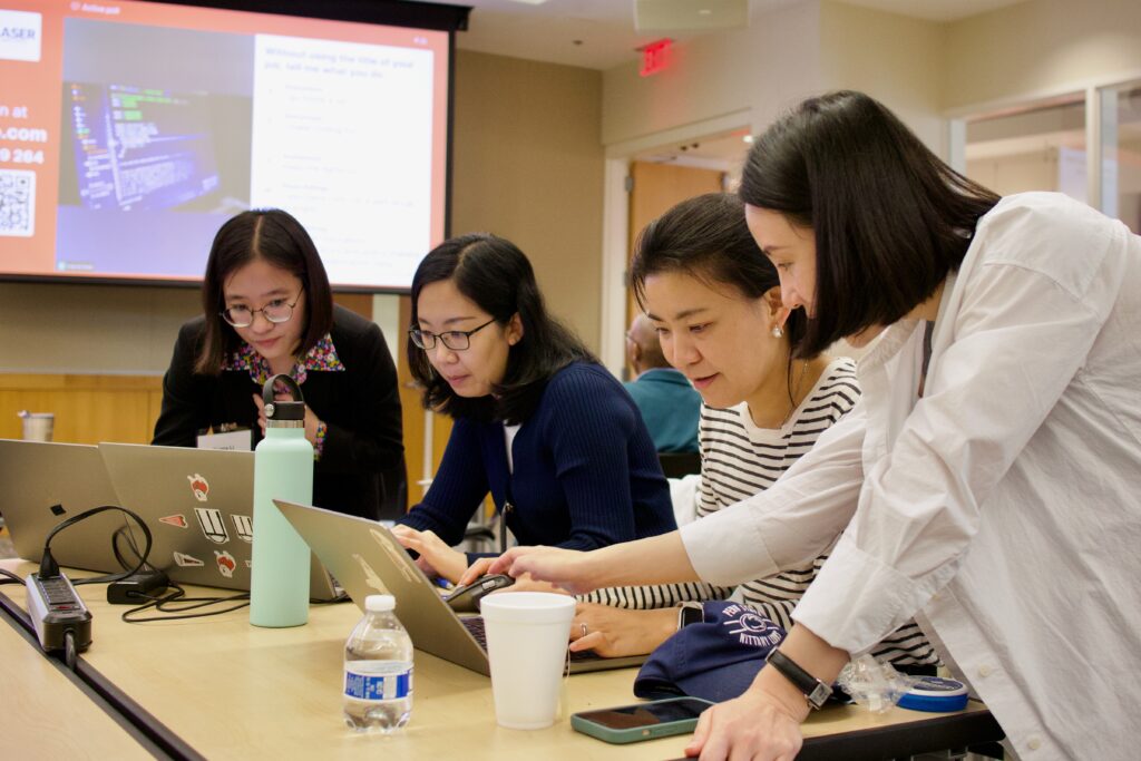 Four women hover over a table talking and pointing at laptops.