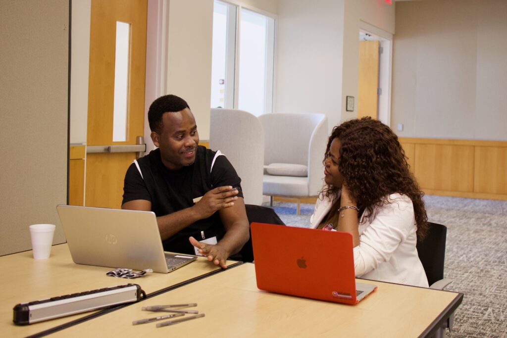 Two people sit at a table in deep conversation. Laptops are in front of them.
