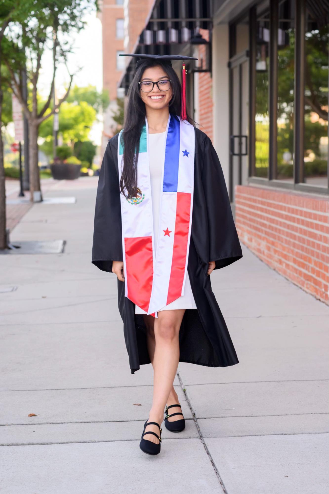 A student in graduation robes and a cap poses on a sidewalk in front of a storefront