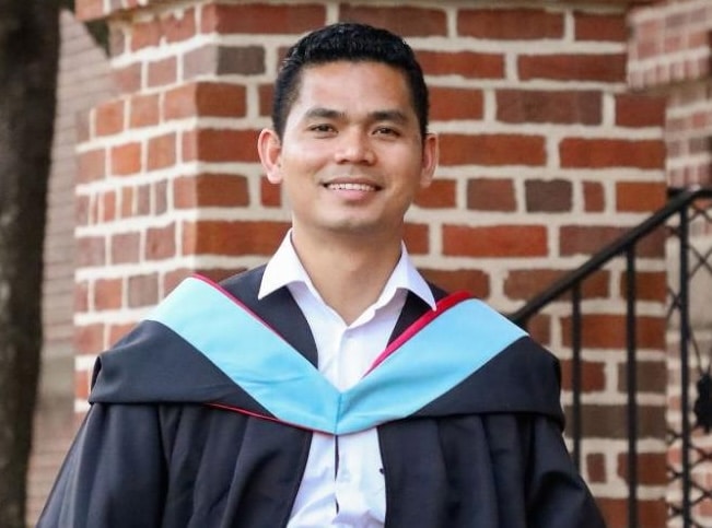 A man in graduation regalia poses in front of a brick building
