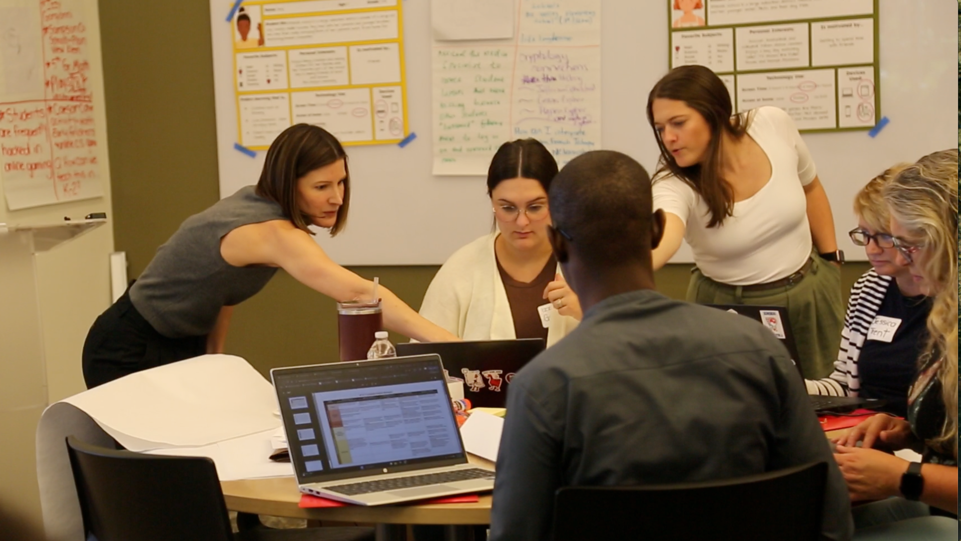 A group of people in a class work around a computer. Some sit at a table while others stand and point to the same laptop screen.