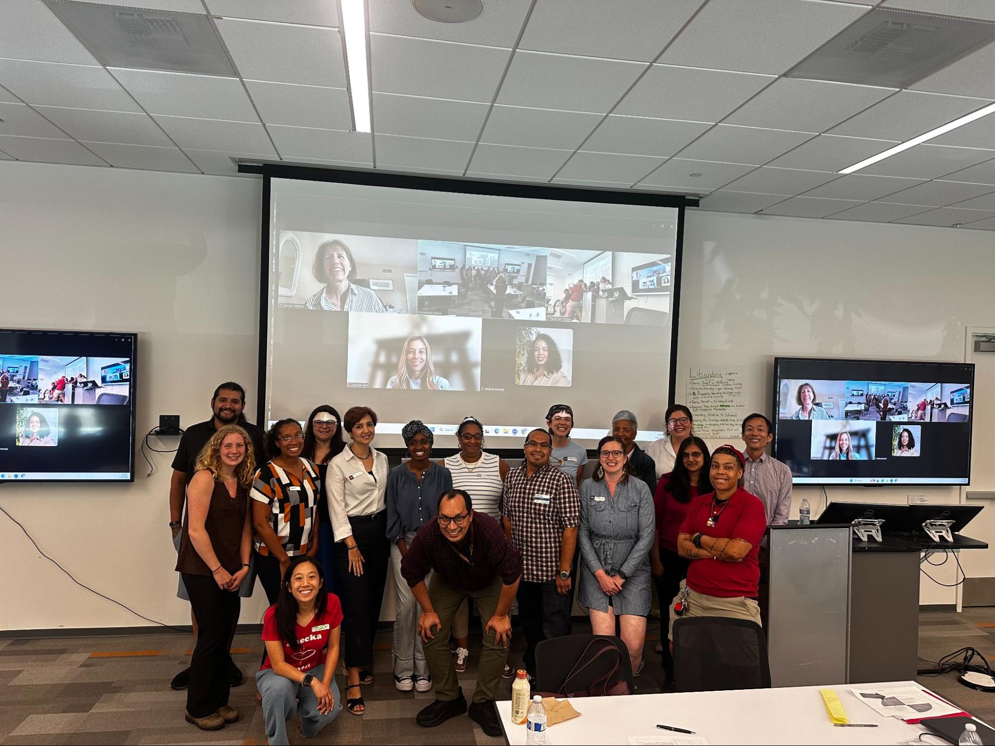 A group of people stand in front of a projector screen filled with a virtual meeting room screen of squares of others