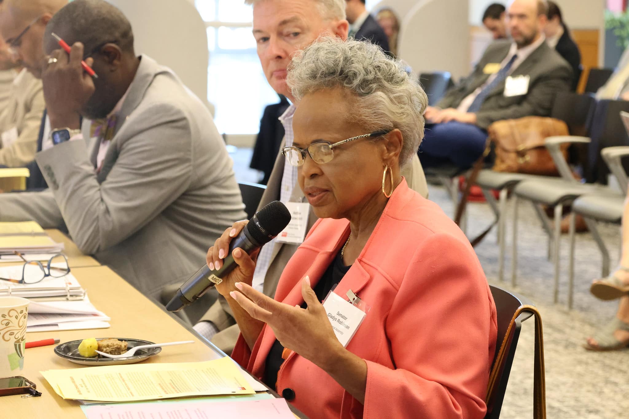 A woman in a salmon blazer speaks on a microphone in a meeting room